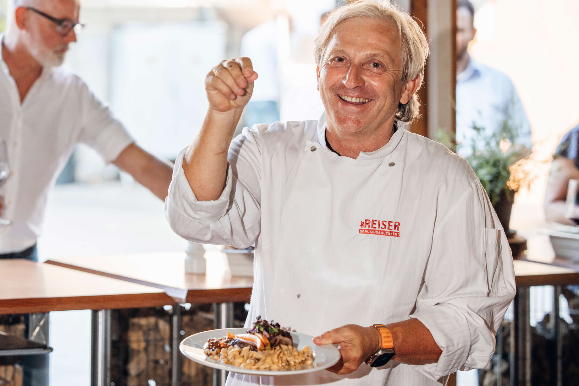 Bernhard Reiser präsentiert kunstvoll dekoriertes Dessert beim Genussfestival, im Hintergrund weitere Gäste.